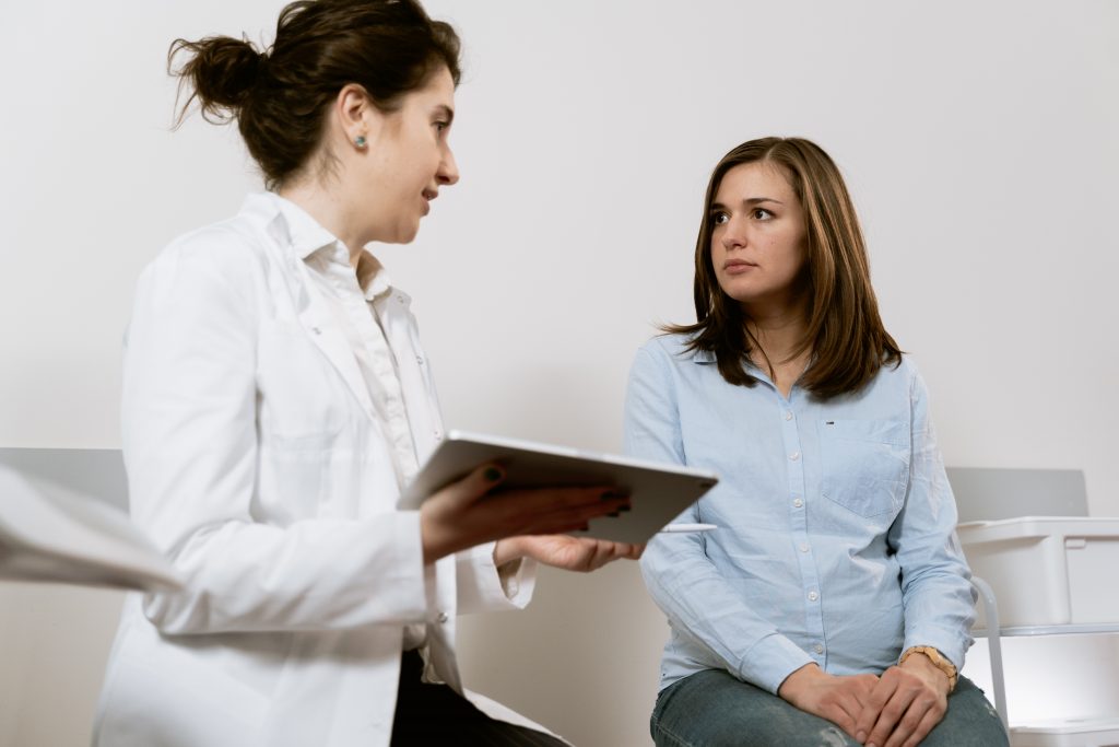 A female physician with a white lab coat and holding a tablet is talking with a female patient who is sitting in front of her